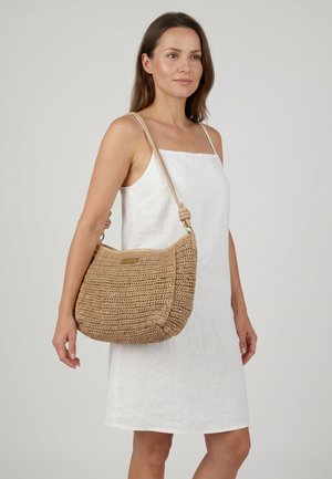 Kvinde i hvid ærmeløs kjole bærer en stor tan vævet skuldertaske med knude strop mod en ensfarvet baggrund.