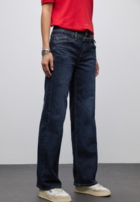 Persona con jeans de piernas anchas de color azul oscuro, zapatillas deportivas blancas, una camisa roja y una pulsera de plata, de pie sobre un suelo gris con un fondo de pared gris claro.