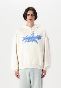 Sudadera oversized color crema con un bolsillo frontal. Presenta un diseño gráfico de estrella azul y una capucha con cordón. Textura de tela suave.