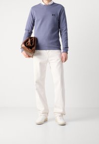 Man som bär en ljusblå tröja, vit skjorta, vita byxor, vita sneakers och håller en brun läderväska mot en vit bakgrund.