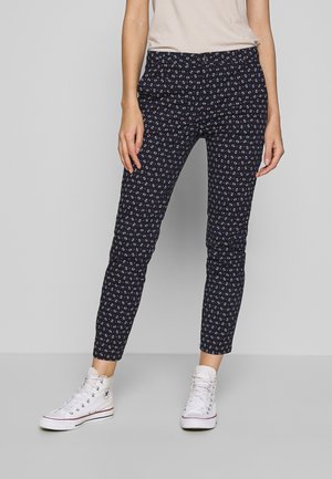Pantalon slim bleu marine avec petits motifs d'ancre blancs et rouges, porté avec des baskets montantes blanches et un haut beige clair.