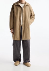 Imperméable léger beige avec boutons-pression, grand col et poches latérales, associé à un pantalon cargo gris et des chaussures beige à enfiler.