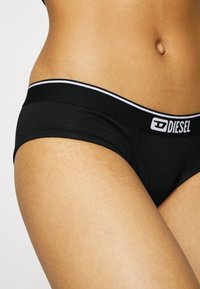Sous-vêtements en coton noir avec une texture en maille, dotés d'une large taille élastique portant le logo "DIESEL" en blanc et d'une fine ligne d'accent blanche.