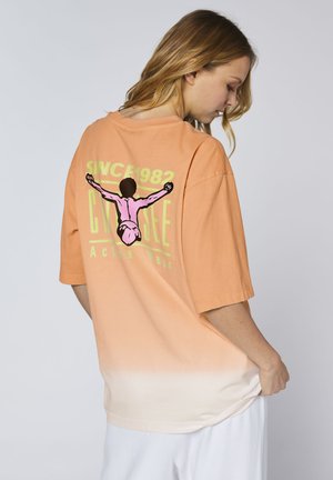 Frau mit langen Haaren, die ein oversized T-Shirt in Orange-Weiß mit Farbverlauf trägt, auf dem eine Grafik einer tauchenden Person und Text auf dem Rücken zu sehen sind.