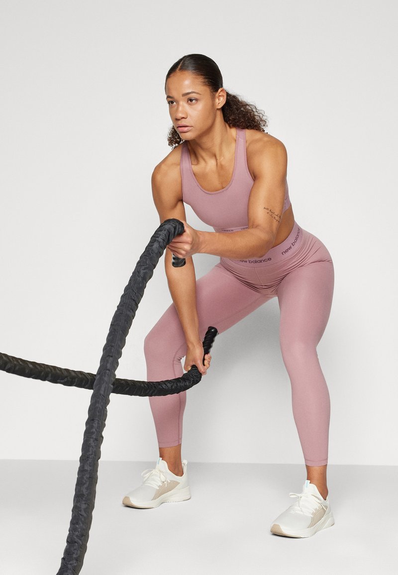 Mujer vestida con ropa deportiva rosa usando cuerdas de batalla en una pose de entrenamiento frente a un fondo liso.