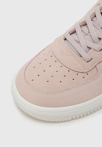 Šviesiai rožiniai zomšiniai sportbačiai su perforuotu viršumi, tekstūruotais sluoksniais ir kreminės spalvos gumine padu. Yra plokščios raišteliai ir logotipo detalė ant liežuvėlio.