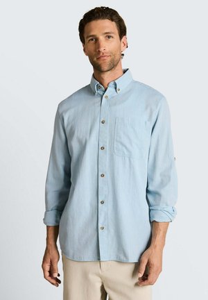 Homme portant une chemise bleu clair à manches longues avec poche poitrine et un pantalon beige, debout devant un fond clair uni.