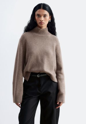 Une femme porte un pull à col roulé côtelé couleur taupe, un pantalon noir et une ceinture sur un fond clair et uni.