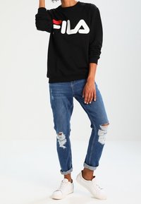 Sudadera negra con logo "FILA" en blanco y rojo, combinada con jeans azules desgastados y zapatillas blancas. Ajuste casual, con puños y dobladillo acanalados.