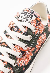 Baskets en toile florales avec des motifs floraux orange et blanc, des feuilles vertes, des lacets blancs, des œillets en argent et un morceau de logo noir.