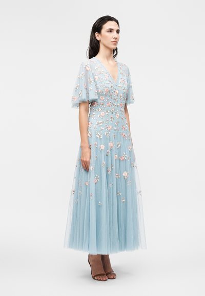 Kvinde iført en lyseblå tyldragt med blomsterbroderi, flagrende ærmer og V-hals, stående på en ensartet hvid baggrund.