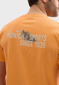Camiseta de algodón naranja con un gráfico de texto gris en la parte posterior: "MÚNICH + DEPORTES DESDE 1939" con una ilustración de zapatillas. Corte casual.