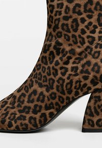 Bottine à imprimé léopard avec un bout pointu, en textile souple, et un talon épais noir. Présente une surface texturée et un motif distinctif.