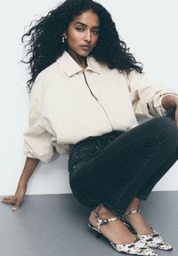 Beige zip-up jasje met ballonmouwen, zwarte slim-fit jeans en witte hakken met een zwart patroon. Model zittend tegen een neutrale achtergrond.