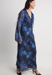 Robe longue en tissu transparent tie-dye bleu, présentant un décolleté en V, des manches longues avec des poignets évasés et un détail de corsage twisté. Talons noirs.