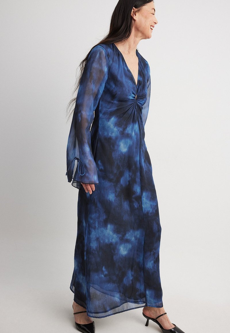 Robe longue en tissu transparent tie-dye bleu, présentant un décolleté en V, des manches longues avec des poignets évasés et un détail de corsage twisté. Talons noirs.