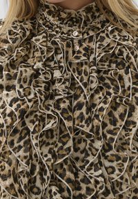 Blouse imprimé léopard avec détails volantés, col montant, tons beige et marron, deux boutons au niveau de l'encolure, tissu texturé.