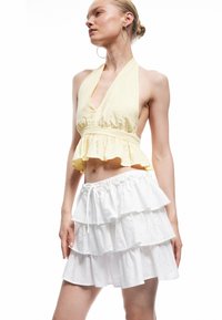 Top corto amarillo con tirantes y dobladillo con volantes, combinado con una falda blanca de capas con patrones florales y cintura con cordón.