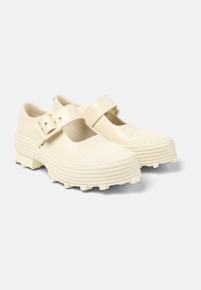 Cream-colored shoes with a rounded toe, zigzag patterns on the rubber sole, and a large adjustable buckle strap across the instep.