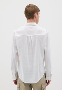 Camicia bianca a maniche lunghe realizzata in tessuto leggero, con yoke posteriore e piega centrale, e polsini abbottonati. Texture liscia, design casual.