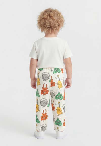 Lille barn med krøllet blondt hår iført hvid t-shirt og cremefarvede bukser med farverige musikinstrument mønstre, står med ryggen til.