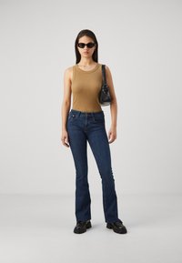 Rjav rebrast top, modre široke kavbojke, črna torbica in temna sončna očala. Model stoji na nevtralni podlagi s straight lasmi.