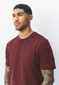 Burgundy cotton t-shirt with short sleeves, crew neck, and a smooth texture, worn by a person against a light background.