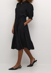 Robe noire avec des boutons sur le devant, manches courtes bouffantes, taille cintrée avec une ceinture et un ourlet évasé. Portée avec des escarpins noirs à bout pointu.