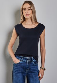 Námořnická modrá bezramínková halenka s kulatým výstřihem, vyrobená z hladkého materiálu. V kombinaci se světle modrými džíny a zlatým náramkem.