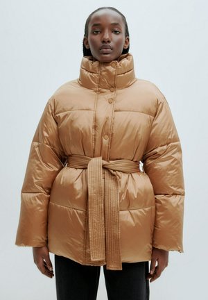 Tan pufferjack met hoge kraag, een ceintuur in de taille en grote gequiltde secties. Zacht, glanzend materiaal en een oversized pasvorm.