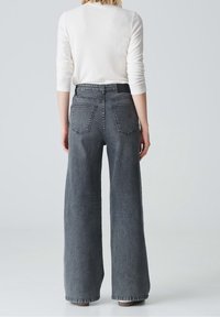 Persona vestida con jeans de tiro alto y pierna ancha de color gris oscuro y una camiseta blanca de manga larga, de pie de espaldas contra un fondo liso.