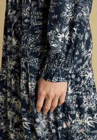 Námornícky kvetinový šaty s bielymi botanickými vzormi, s naťahovacími zápästnými časťami a splývavou siluetou. Viditeľná je jemná textúra látky.