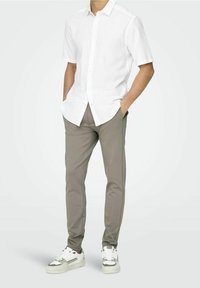 Man in een witte korte mouwen overhemd met knopen, grijze slim-fit broek en witte sneakers met grijze details, handen in de zakken.