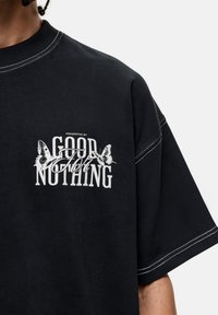 T-shirt noir oversize en coton, avec un texte blanc et des graphismes de papillon sur la poitrine. Des coutures contrastées soulignent les manches.