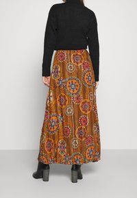 Gonna maxi marrone con motivi circolari colorati. Presenta un design plissettato e una forma fluida, abbinata a un top nero e stivali con tacco.
