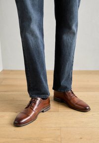 Brown leather dress shoes with a rounded toe, lace-up front, and a stitched sole. Paired with dark denim jeans on a wooden floor.