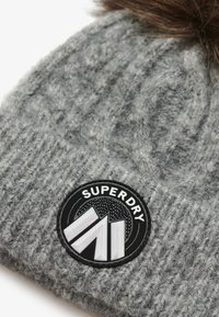 Gorro de punto gris con un acabado texturizado y un pompón de piel marrón. Presenta un parche circular negro con el texto "SUPERDRY" y un diseño geométrico.