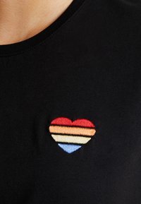 Hjerteformet broderi med røde, orange, cremede og blå vandrette striber på et sort stof, placeret nær brystområdet.