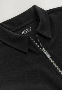 Black zip-up polo shirt with a textured fabric, featuring a collar and silver zipper. Tag reads "NEXT," indicating manufacture in Bangladesh.