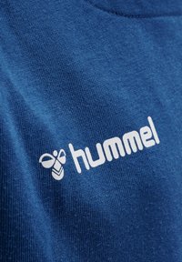 Blauwe stof met wit hummel-logo en bijensymbool gedrukt op het gestructureerde oppervlak van een kledingstuk.
