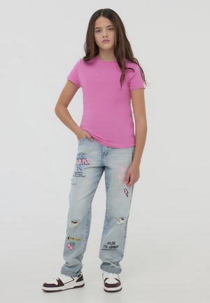 T-shirt rosa aderente, jeans chiari strappati con toppe colorate e ricami, sneakers bianche e marroni, in posa in piedi.