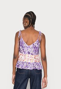 Donna con i capelli intrecciati che indossa orecchini a cerchio e un top senza maniche viola con una stampa floreale e spalline annodate, mostrata di spalle.