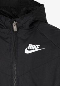 Musta Nike-takki vetoketjulla, jossa valkoinen Nike-logo rinnassa. Materiaali on sileä; suunnittelussa on huppu ja ompelut.