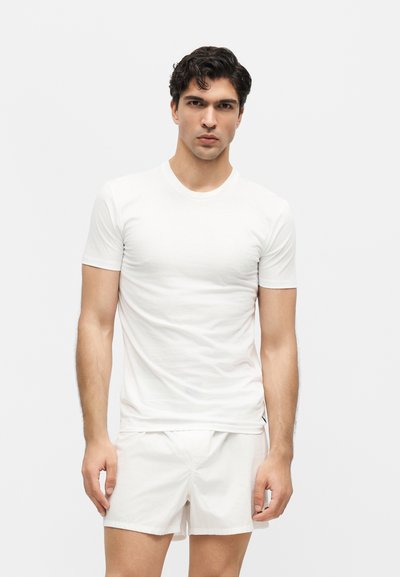 Νέος με σκούρα μαλλιά, που φοράει απλό λευκό κοντομάνικο μπλουζάκι και λευκά σορτς, στέκεται μπροστά σε ένα απλό φωτεινό φόντο.