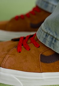 Zapatillas de ante marrón con cordones rojos, presentando un acento marrón oscuro y suela de goma blanca. Combinadas con jeans de color azul claro, que muestran superficies texturizadas.