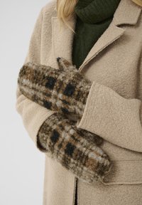 Brune og creme ternede vanter lavet af tekstureret uld, vist hvilende mod en beige frakke og en grøn sweater. Vanterne har et tommel-design.