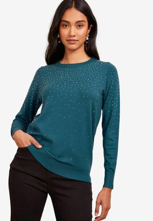 Teal Pullover mit langen Ärmeln, rundem Halsausschnitt und verstreuten Strassverzierungen. Getragen mit schwarzen Hosen, die eine lockere Passform haben.