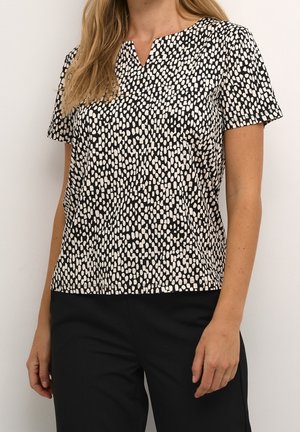 Femme vêtue d'un haut à manches courtes à motif noir et blanc avec un pantalon noir, debout devant un fond blanc uni.