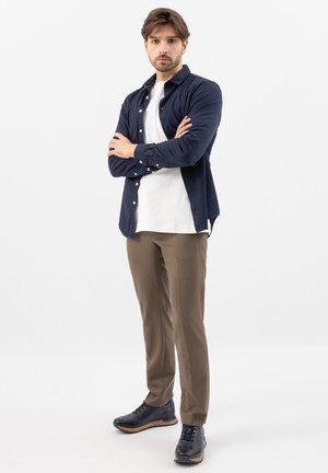 Jonge man met gevouwen armen, gekleed in een marineblauw overhemd, wit T-shirt, bruine broek en zwarte sneakers, staand tegen een witte achtergrond.