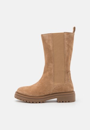 Botte en suède beige, hauteur genoux, avec des panneaux latéraux en élasthanne, bout rond, semelle texturée et perforations décoratives près du talon.